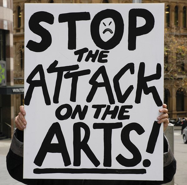 Black text on white placard says Stop the Attack on the Arts. There are squiggly lines and a sad face in the letters.