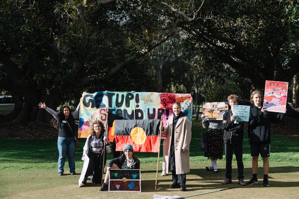 Seven teenagers stand in front of trees holding up protest banners, with one reading get up! stand up! and another being a painted Aboriginal flag.