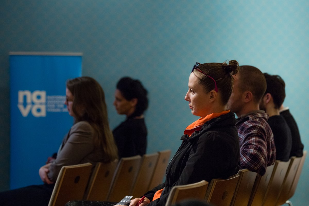 Photo of people sitting in rows and looking forward away from the camera as though listening to someone out of shot. In the background is a white NAVA logo on a blue background.