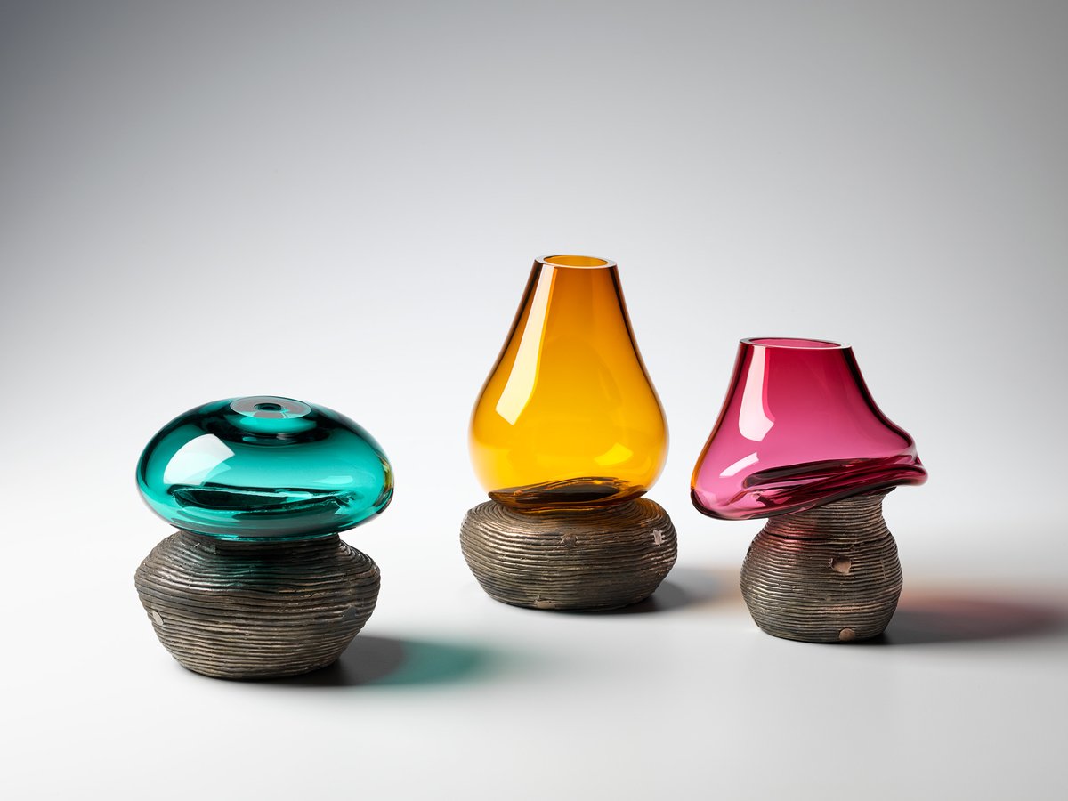 A photo of three jewel-toned glass vessels, each resting on top of a brass vessel. The glass is organically shaped and appears to slump into the surface of the brass. The three objects are against a plain white background.