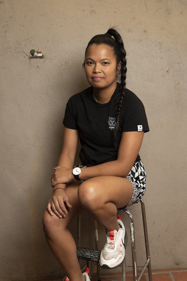 Image of a woman sitting on a stool against a brown backdrop. She has black hair that is pulled back into a long braided pony tail. She is wearing a black tshirt, black and white patterned shorts, white sneakers and a watch with a black strap. 