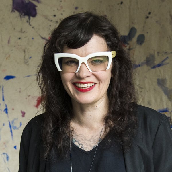 Head and shoulders portrait of Penelope Benton. She has brown wavy hair, white framed glasses, red lipstick and is wearing a black top. In the background is a canvas of paint splash marks.