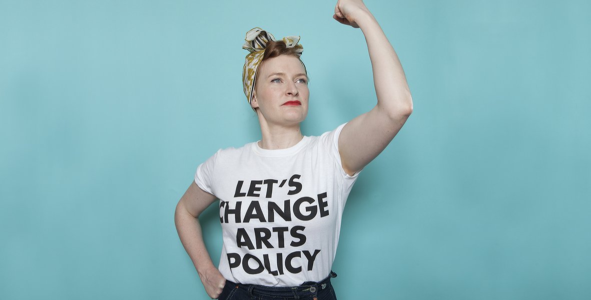 A white woman standing in front of a turquoise background wearing a white shirt with the text in black 'Lets Change Arts Policy'. She has one hand on her hip and one hand above her head in a fist.
